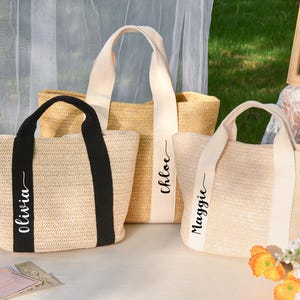 K&ouml;nnte beinhalten: Drei gewebte Stroh-Tragetaschen mit unterschiedlichen Griffen. Eine Tasche hat schwarze Griffe und den Namen "Olivia" aufgedruckt. Eine andere hat cremefarbene Griffe mit dem Namen "Chloe". Die dritte Tasche hat cremefarbene Griffe mit dem Namen "Maggie".