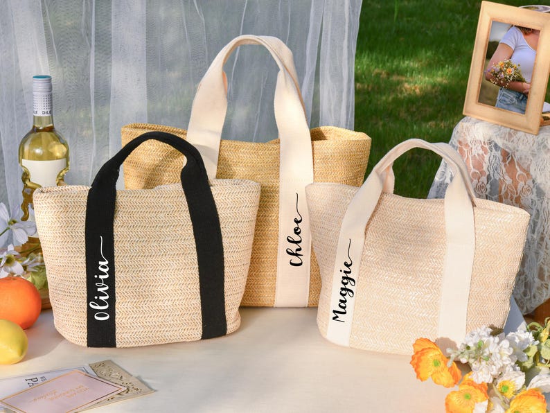K&ouml;nnte beinhalten: Drei gewebte Stroh-Tragetaschen mit unterschiedlichen Griffen. Eine Tasche hat schwarze Griffe und den Namen "Olivia" aufgedruckt. Eine andere hat cremefarbene Griffe mit dem Namen "Chloe". Die dritte Tasche hat cremefarbene Griffe mit dem Namen "Maggie".
