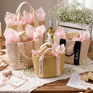 K&ouml;nnte beinhalten: Vier gewebte Stroh-Tragetaschen mit rosa Seidenpapier, Geschenken und personalisierten B&auml;ndern. Die Taschen haben cremefarbene Griffe und die Namen "Chloe", "Evelyn" und "Ashley" sind auf den B&auml;ndern aufgedruckt. Eine Weinflasche und Blumen sind ebenfalls vorhanden.