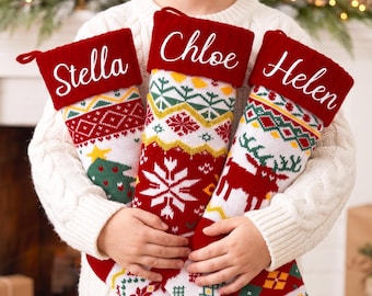 May include: Three knitted Christmas stockings featuring red cuffs with the white embroidered names Stella, Chloe, and Helen. The stockings display a festive pattern of white, red, and green geometric shapes, snowflakes, a Christmas tree, and a reindeer.