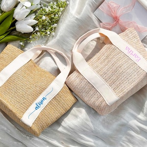 K&ouml;nnte beinhalten: Zwei gewebte Tragetaschen mit wei&szlig;en Tr&auml;gern und personalisiertem Text. Eine Tasche ist naturfarben mit "alison" in blauer Schrift, die andere ist rosa mit "Eartha" in rosa Schrift. Die Taschen liegen auf einer wei&szlig;en, strukturierten Oberfl&auml;che.