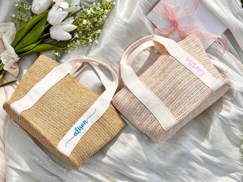 K&ouml;nnte beinhalten: Zwei gewebte Tragetaschen mit wei&szlig;en Tr&auml;gern und personalisiertem Text. Eine Tasche ist naturfarben mit "alison" in blauer Schrift, die andere ist rosa mit "Eartha" in rosa Schrift. Die Taschen liegen auf einer wei&szlig;en, strukturierten Oberfl&auml;che.