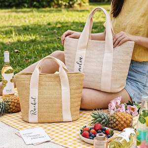 K&ouml;nnte beinhalten: Zwei gewebte Stroh-Tragetaschen mit cremefarbenen Griffen und Namensschildern. Auf einer Tasche steht "Rachel" und auf der anderen "Amelia". Die Taschen liegen auf einer gelb-wei&szlig; karierten Picknickdecke mit Obst, Getr&auml;nken und Blumen.