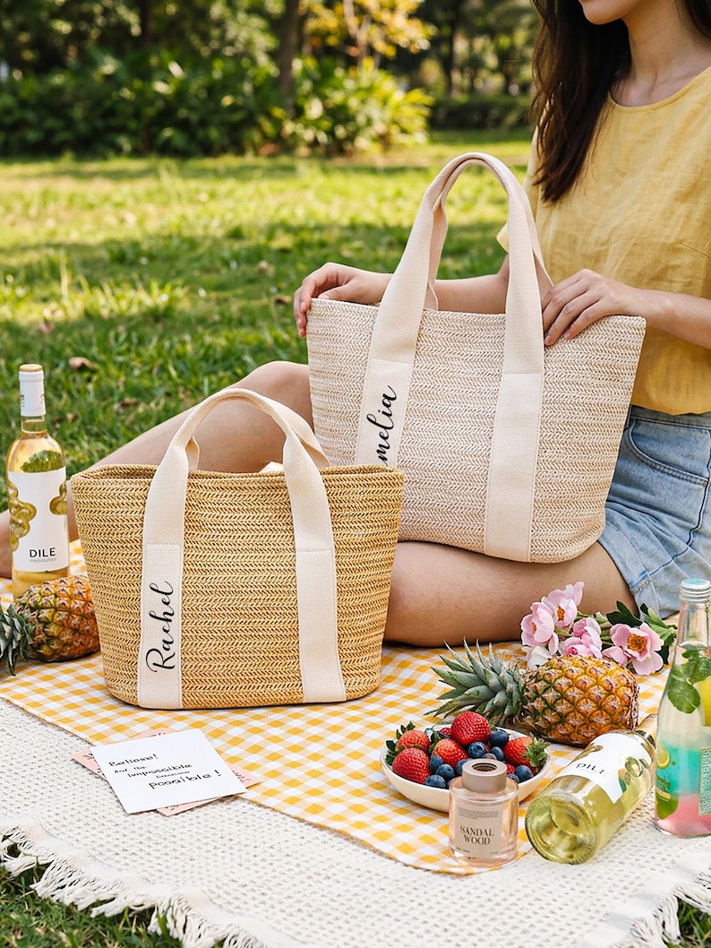 K&ouml;nnte beinhalten: Zwei gewebte Stroh-Tragetaschen mit cremefarbenen Griffen und Namensschildern. Auf einer Tasche steht "Rachel" und auf der anderen "Amelia". Die Taschen liegen auf einer gelb-wei&szlig; karierten Picknickdecke mit Obst, Getr&auml;nken und Blumen.