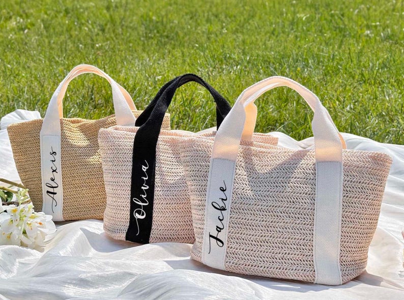 K&ouml;nnte beinhalten: Drei gewebte Tragetaschen mit Griffen, jede mit einem anderen Farbschema. Eine ist naturfarben, eine rosa und eine schwarz. Jede Tasche hat ein wei&szlig;es Band mit einem Namen in Schreibschrift: Alexis, Olivia und Jackie. Die Taschen liegen auf einem wei&szlig;en Tuch.