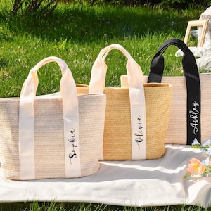 K&ouml;nnte beinhalten: Drei gewebte Tragetaschen mit personalisierten Namen: Sophia, Olivia und Ashley. Die Taschen haben cremefarbene K&ouml;rper mit passenden oder schwarzen Griffen und Namensb&auml;ndern. Die Szene spielt sich im Freien auf einer wei&szlig;en Decke mit Blumen und einer Weinflasche ab.