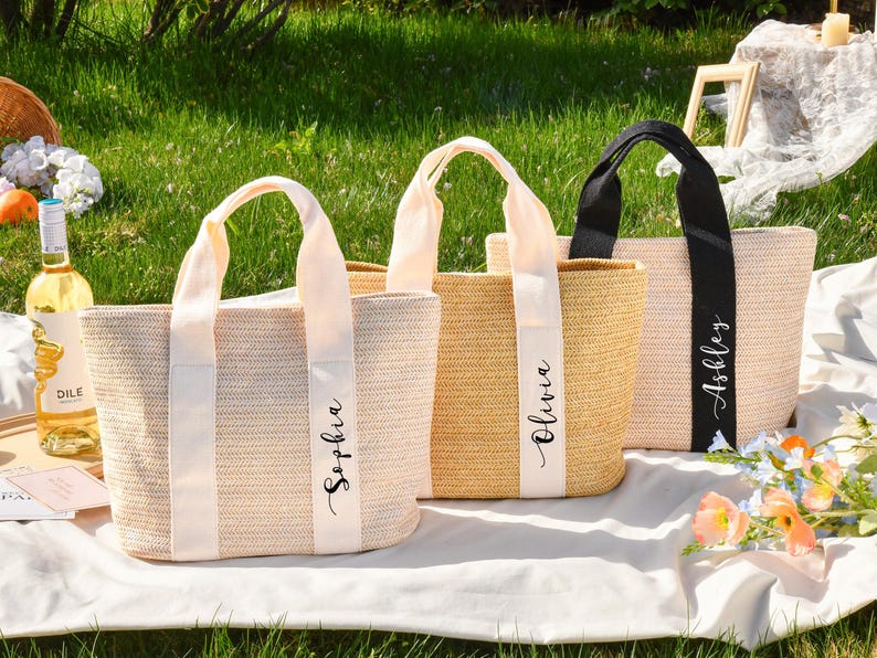 K&ouml;nnte beinhalten: Drei gewebte Tragetaschen mit personalisierten Namen: Sophia, Olivia und Ashley. Die Taschen haben cremefarbene K&ouml;rper mit passenden oder schwarzen Griffen und Namensb&auml;ndern. Die Szene spielt sich im Freien auf einer wei&szlig;en Decke mit Blumen und einer Weinflasche ab.