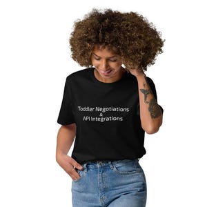 May include: A black t-shirt with the text "Toddler Negotiations & API Integrations" in white, paired with light blue jeans and white sneakers. The person is standing against a white background.