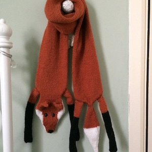 May include: A knitted fox scarf in orange, black, and white. The scarf has a fox head with black eyes and a white nose. The scarf has two long tails that are black and white.