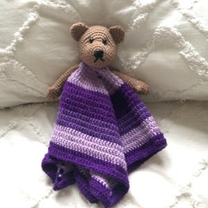 May include: A crocheted purple and lavender striped baby blanket with a brown crocheted teddy bear attached.