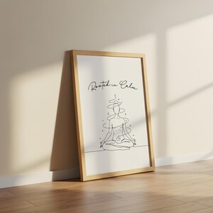 May include: A framed art print featuring a minimalist line drawing of a person in a seated yoga pose. The artwork is in black and white, with the words "Rooted in Calm" in script at the top. The frame is light brown.