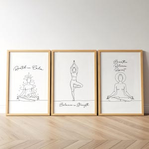 May include: Three framed art prints with line drawings of yoga poses. The prints have a white background and a natural wood frame. Text includes "Rooted in Calm", "Balance is Strength", and "Breathe Release Repeat".