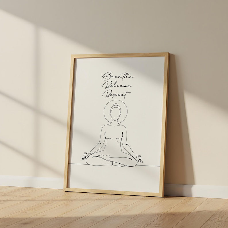 May include: A framed art print featuring a minimalist line drawing of a person in a seated yoga pose. The artwork is in black and white, with the words "Breathe Release Repeat" in elegant script above the figure. The frame is a light wood color.