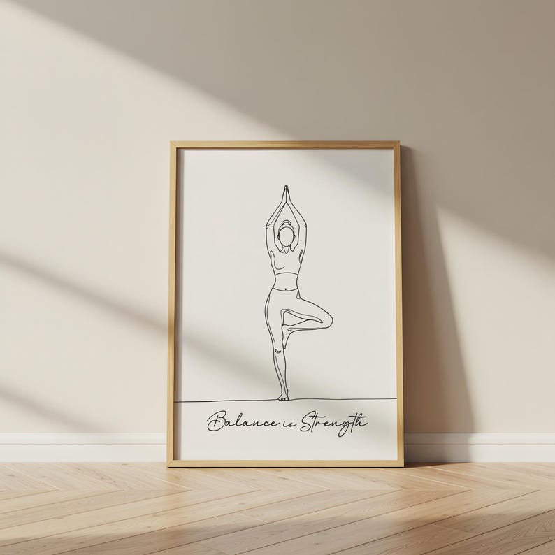 May include: A framed print featuring a minimalist line drawing of a person in a yoga tree pose. The artwork is in black against a cream background, with the words "Balance is Strength" in script below the figure. The frame is a light wood color.