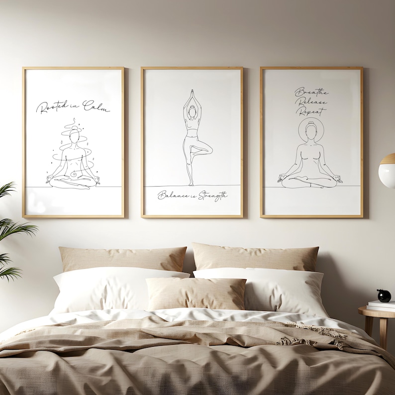 May include: Three framed art prints with line drawings of yoga poses and inspirational text. The prints are in light wood frames and hang above a bed with neutral-toned bedding. The text includes "Rooted in Calm", "Balance is Strength", and "Breathe, Release, Repeat."