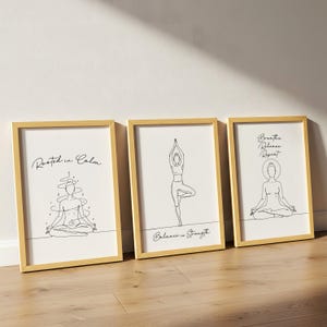 May include: Three framed art prints with gold-colored frames. Each print features a minimalist line drawing of a yoga pose and inspirational text. The prints are displayed on a light wood floor.
