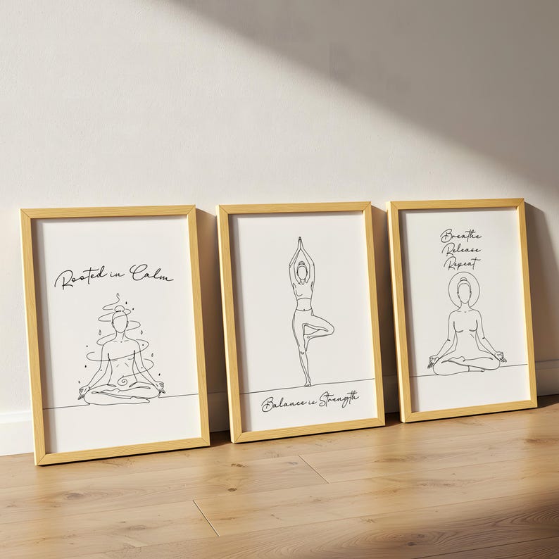 May include: Three framed art prints with gold-colored frames. Each print features a minimalist line drawing of a yoga pose and inspirational text. The prints are displayed on a light wood floor.