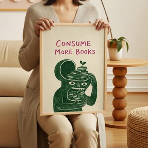 Puede incluir: Un cuadro enmarcado con las palabras "CONSUME MORE BOOKS" en rosa. El cuadro presenta una ilustraci&oacute;n verde de una figura con libros en la cabeza y la boca. El marco es de color madera clara.