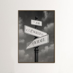 May include: Black and white framed print featuring street signs with the names "Angelina" and "Michael." The top sign reads "EST 2018." The background is a cloudy sky.