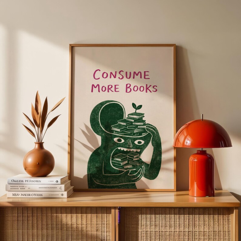 Puede incluir: Una impresi&oacute;n art&iacute;stica enmarcada con el texto "CONSUME MORE BOOKS" sobre una figura de dibujos animados verde que sostiene y come libros. Una l&aacute;mpara de mesa roja y un jarr&oacute;n marr&oacute;n con plantas secas est&aacute;n sobre un mueble de madera.