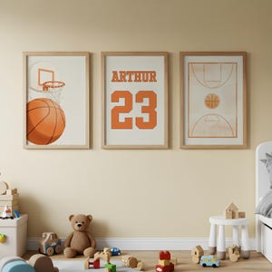 Könnte beinhalten: Drei gerahmte Basketball-Prints. Einer zeigt einen Basketball und einen Korb, ein anderer zeigt den Namen "ARTHUR" und die Nummer "23", und der dritte zeigt ein Basketballfeld. Die Prints haben einen hellen Holzrahmen und werden an einer cremefarbenen Wand gezeigt.