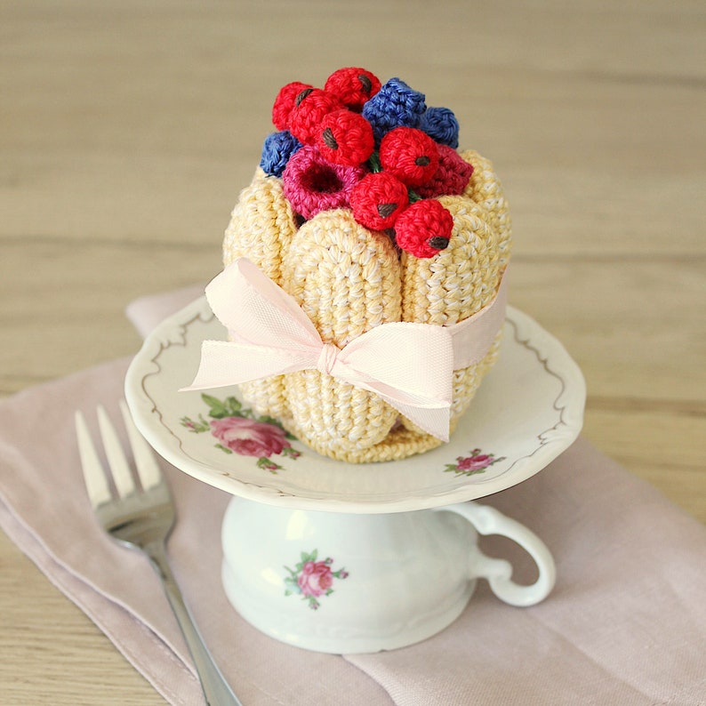 Puede incluir: Un pastel de crochet con una cinta rosa y una cobertura de bayas rojas y azules, presentado en una taza de t&eacute; y un platillo blancos con dise&ntilde;os florales.
