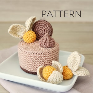 May include: A crocheted cake with decorative elements, including a brown base, a light pink body, and various toppings. The toppings include a brown spiral, a small brown teardrop, and yellow and cream-colored accents. The word "PATTERN" is displayed.