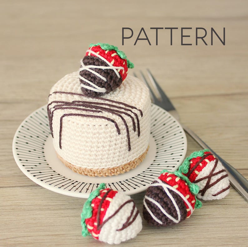 A crocheted mini cheesecake sits on a plate. It has got a cookie crumble cake base with two shades of brown. This amigurumi cake is topped with a chocolate covered strawberry. Everything is made out of cotton yarn.