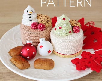 May include: A festive holiday display featuring crocheted miniature cakes topped with a snowman and Christmas tree. The cakes sit on a white plate with red and white ornaments, gingerbread cookies, and red felt decorations. The word "PATTERN" is displayed in white text.