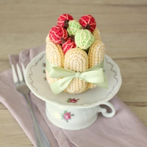 Puede incluir: Una r&eacute;plica de postre de ganchillo con una base amarilla y una cobertura de fruta verde y roja. El postre est&aacute; atado con una cinta verde y est&aacute; sentado en un platillo blanco con dise&ntilde;os florales.