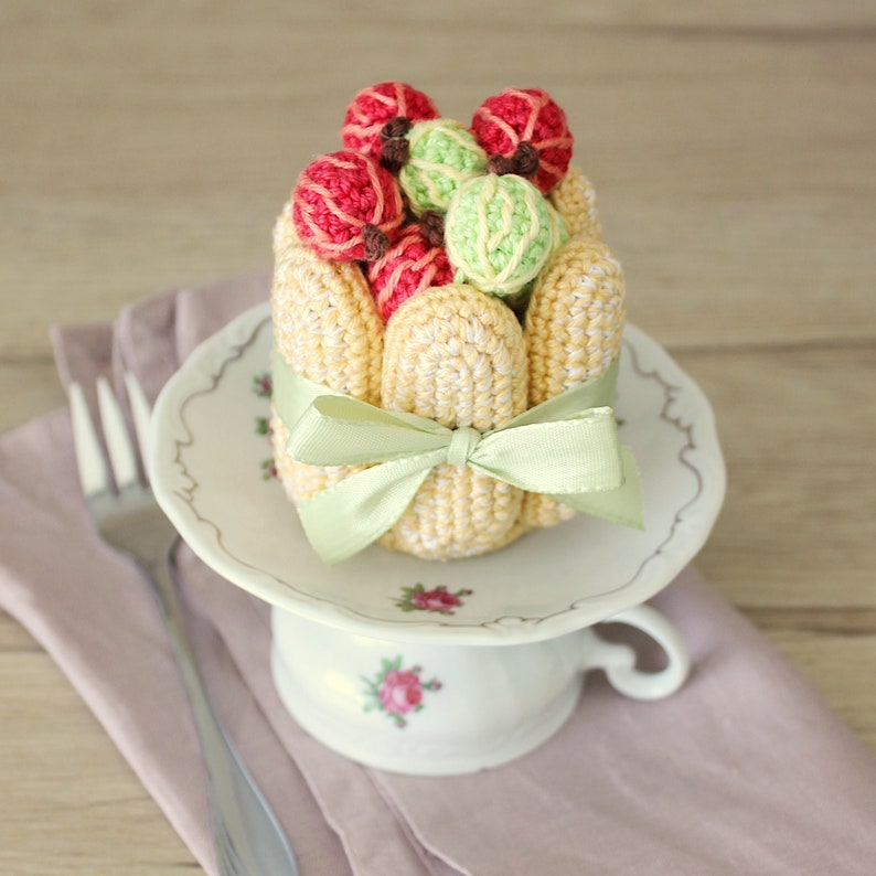 Puede incluir: Una r&eacute;plica de postre de ganchillo con una base amarilla y una cobertura de fruta verde y roja. El postre est&aacute; atado con una cinta verde y est&aacute; sentado en un platillo blanco con dise&ntilde;os florales.