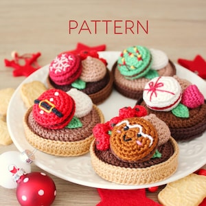 Op de afbeelding: Een witte plaat toont verschillende gehaakte kerstdesserts, elk met unieke toppings zoals een kerstman, sneeuwvlok en peperkoekman. Het woord "PATTERN" staat in het rood boven de plaat. Ornamenten en koekjes omringen de plaat.