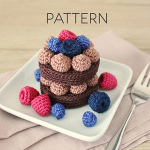 May include: A crocheted dessert with a dark brown base, adorned with pink, blue, and tan yarn berries. The word "PATTERN" is displayed above the dessert. The dessert sits on a white square plate with a fork and a pink napkin.
