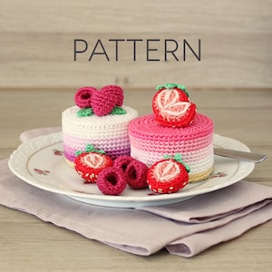 Op de afbeelding: Gehaakte miniatuur taarten met aardbeien- en frambozentoppings, gepresenteerd op een decoratief bord. De taarten hebben roze, witte en paarse kleurovergangen. Het woord "PATTERN" wordt boven de taarten weergegeven.