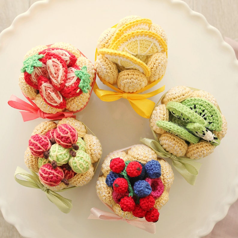 Puede incluir: Cinco cupcakes de ganchillo con tem&aacute;tica de frutas con detalles amarillos, verdes y rojos. Los cupcakes est&aacute;n dispuestos en c&iacute;rculo sobre un plato blanco.