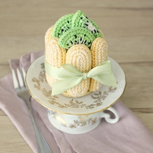 Puede incluir: Un postre de kiwi tejido a crochet con un lazo verde sobre una taza de t&eacute; y un platillo blancos.