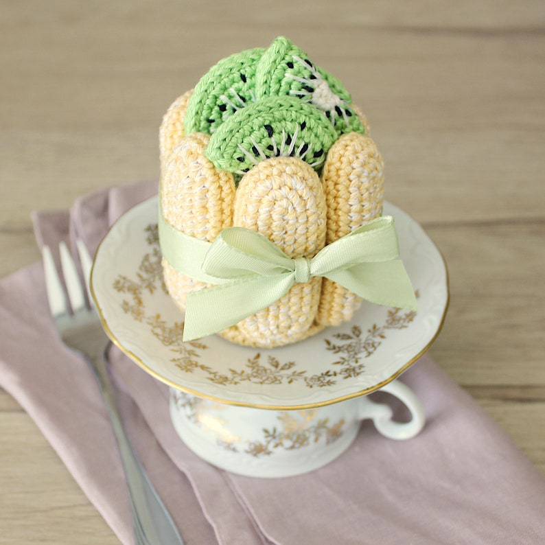 Puede incluir: Un postre de kiwi tejido a crochet con un lazo verde sobre una taza de t&eacute; y un platillo blancos.