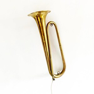 Pode incluir: Uma trombeta de latão vintage com sino alargado e tubos curvos. O instrumento tem uma cor dourada quente e parece estar montado numa superfície branca. A trombeta tem um design clássico, sugerindo que pode ser um item de colecionador ou decorativo.