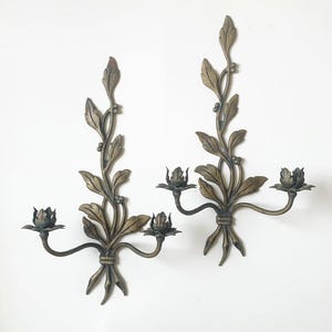 May include: A pair of wall-mounted candle sconces crafted from dark bronze-toned metal. Each piece features a central vertical stem adorned with sculpted leaves and small berries, branching into two curved arms that hold flower-shaped candle cups.