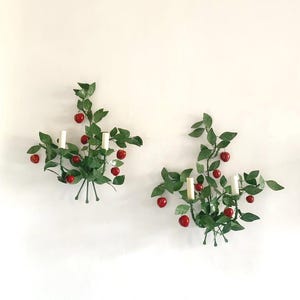 Puede incluir: Dos apliques de pared con diseño botánico. Cada aplique presenta hojas verdes, detalles rojos en forma de cereza y dos candelabros blancos. Los apliques están montados en una pared blanca.