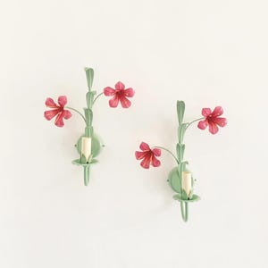 Peut inclure: Deux appliques murales florales. Chaque applique présente une base vert clair avec une tige et trois fleurs rouges. Les appliques sont fixées sur un mur blanc, ajoutant une touche de nature à la décoration.
