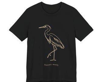 May include: A black short-sleeve t-shirt featuring a gold-colored line art illustration of a standing heron. Below the bird, the text "Patience, mostly." is printed in a small, simple, light-colored serif font centered on the chest.