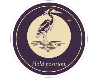 May include: A circular sticker featuring a dark purple border and a cream-colored center. The design displays a black heron standing in water, a small orange four-pointed star, and the text Hold position. in a serif font at the bottom.
