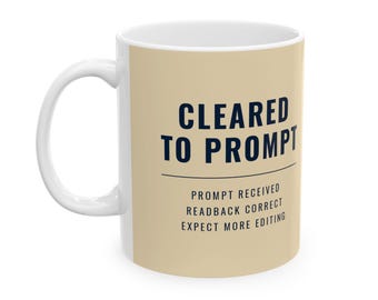 May include: A beige ceramic mug with a white interior and handle. The front features dark blue text that reads CLEARED TO PROMPT, followed by a horizontal line and the text PROMPT RECEIVED, READBACK CORRECT, EXPECT MORE EDITING.