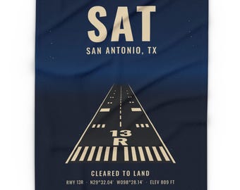May include: A dark blue blanket featuring a graphic of an airport runway. The text reads SAT, SAN ANTONIO, TX, 13 R, CLEARED TO LAND, RWY 13R, N29°32.04' W098°28.14', and ELEV 809 FT. The design includes white runway markings on a dark background.