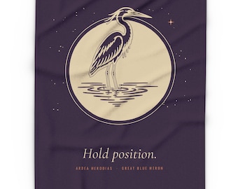 May include: A dark purple blanket featuring a cream-colored circular graphic of a great blue heron standing in water. The design includes small stars and the text "Hold position." followed by "ARDEA HERODIAS - GREAT BLUE HERON" below.
