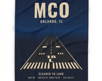 May include: A dark blue blanket featuring a graphic of an airport runway. The text reads MCO, ORLANDO, FL, CLEARED TO LAND, RWY 18L, N28°25.76' W081°18.54', and ELEV 96.3 FT. The design includes a perspective view of the runway markings and numbers.