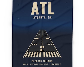 May include: A dark blue fabric blanket featuring a graphic of an airport runway. The text reads ATL, ATLANTA, GA, CLEARED TO LAND, RWY 8L, N33°38.20' W084°25.67', and ELEV 1026.2 FT in cream-colored lettering against the dark background.
