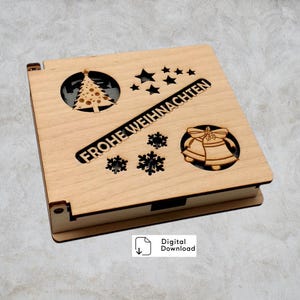 May include: A light-coloured wooden box with laser-cut Christmas-themed designs. The lid features a Christmas tree, stars, snowflakes, bells, and the text "FROHE WEIHNACHTEN". The box is rectangular and has a hinged lid.