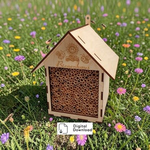 Può includere: Una casetta di legno per api con tetto a capanna, riempita di tubi di bambù. La casa è incisa con un girasole, api e la parola "Bienenhaus". L'immagine è ambientata in un campo di fiori selvatici colorati.